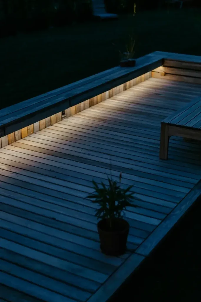 A dimly lit wooden deck with built-in bench seating, subtle lighting, and potted plants at night.