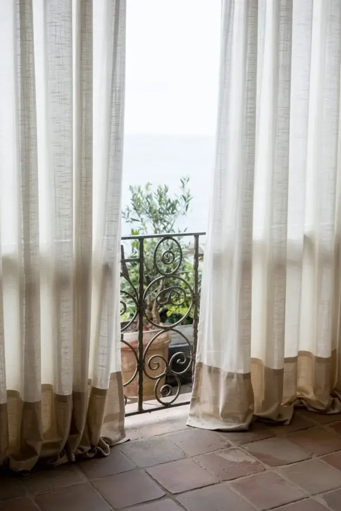 Sheer curtains framing a wrought iron balcony overlooking a sea view.