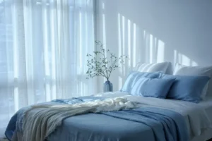Sunlit bedroom with blue and white bedding, sheer curtains, and a vase with branches casting shadows on the wall.