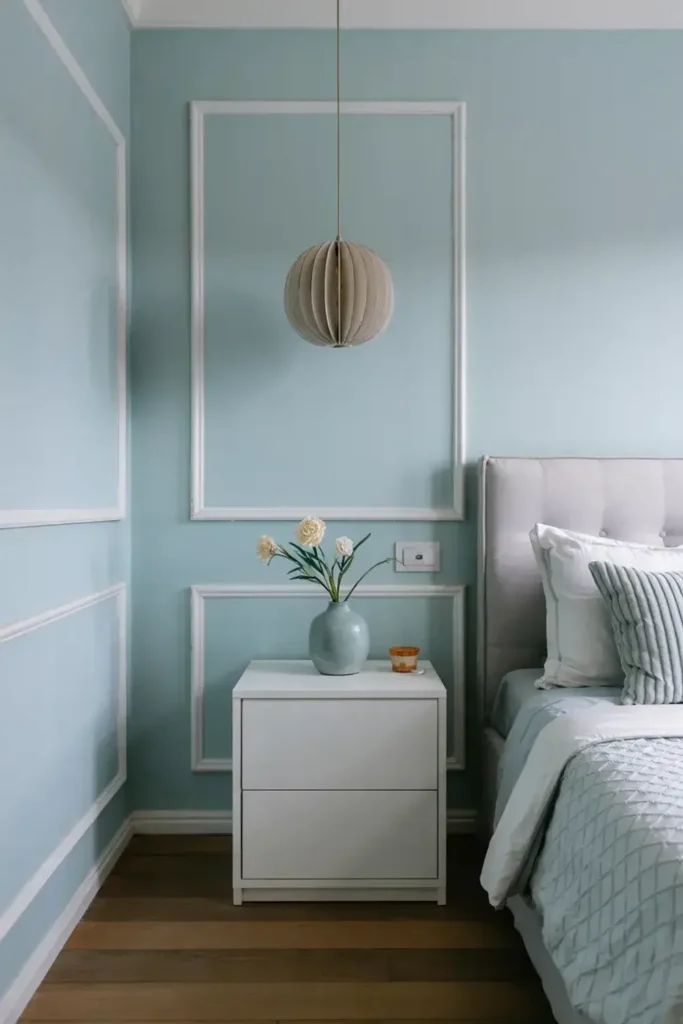 Light blue bedroom corner with white molding, nightstand, vase with flowers, pendant light, and grey bed.