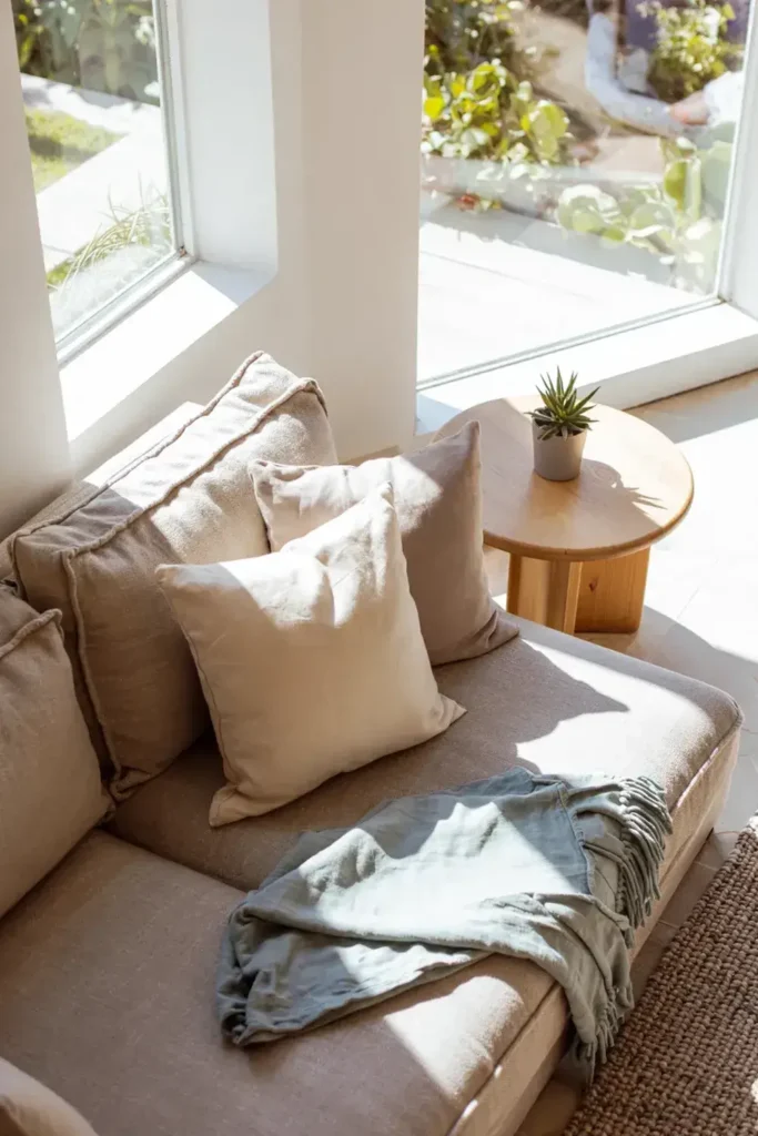 Sunlit reading nook with cushions, throw blanket, round wooden side table, and small potted succulent by expansive windows.