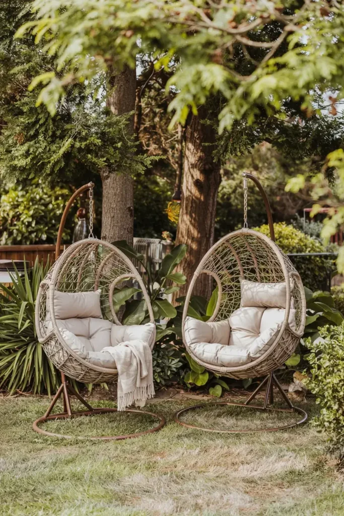 Two wicker hanging egg chairs with cream cushions sit in a lush green garden.