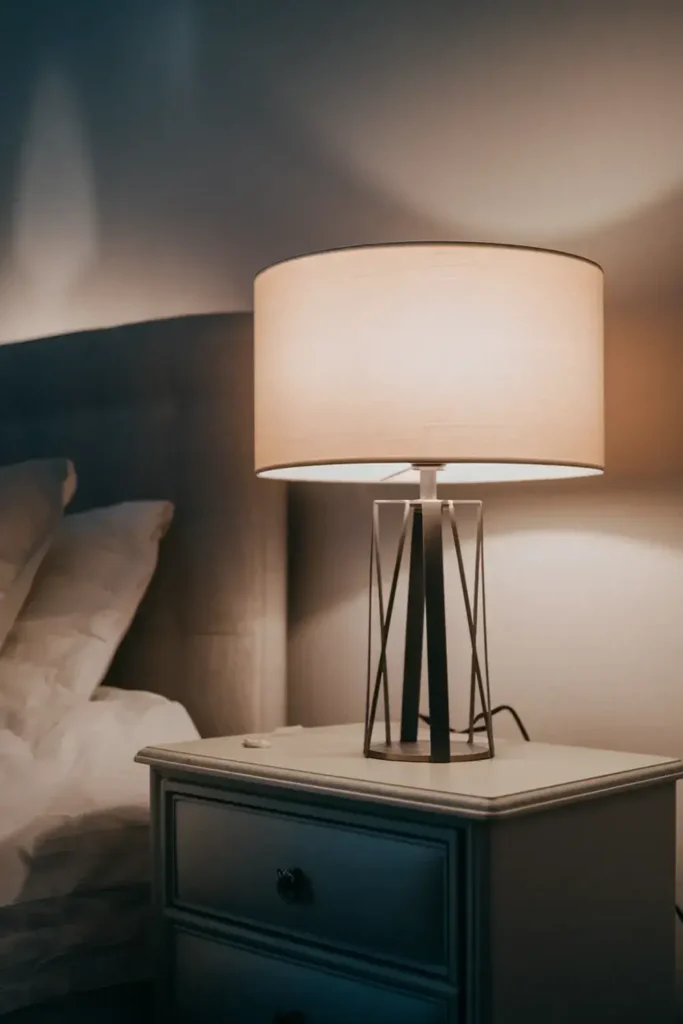 Illuminated bedside table lamp with geometric base on a nightstand next to a bed.